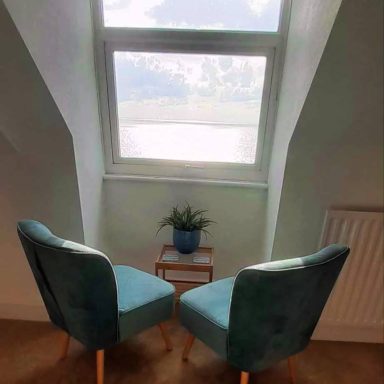 Two blue chairs , a small table with a plant. Infront of a window with sea view.